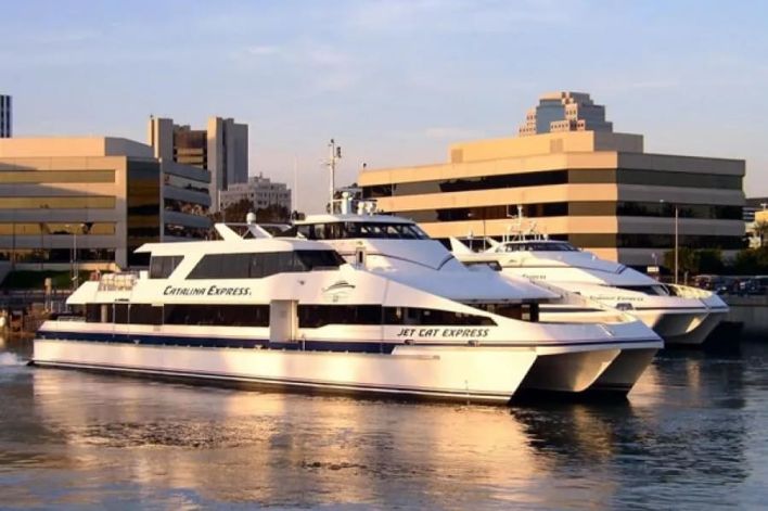 Two passenger ferries, named Catalina Express and Jet Cat Express, are docked side by side in a harbor near modern office buildings at sunset.