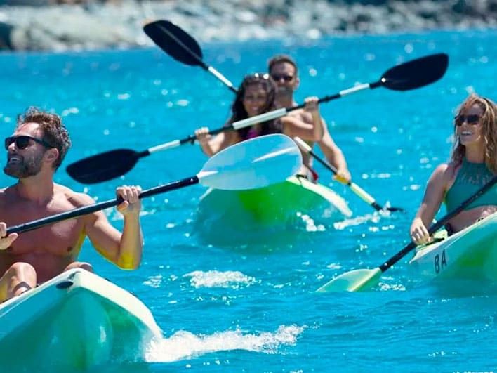 Four people paddle two green kayaks on bright blue water, with a rocky shoreline visible in the background.