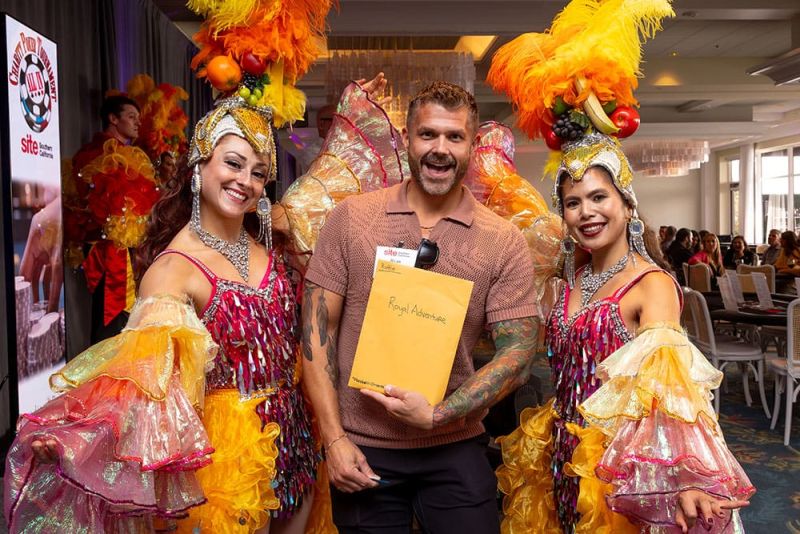 A man holding a yellow envelope labeled "raffle - Royal Adventure" poses between two women in colorful carnival costumes with feathered headpieces at the SITE SoCal Poker Tournament..